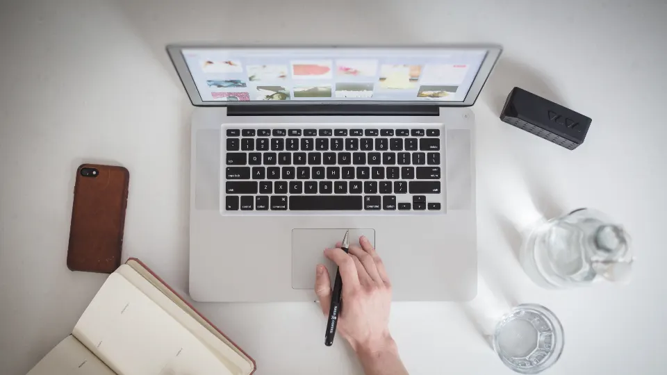 Vista superior de una persona trabajando en un portátil con libreta y celular sobre un escritorio blanco
